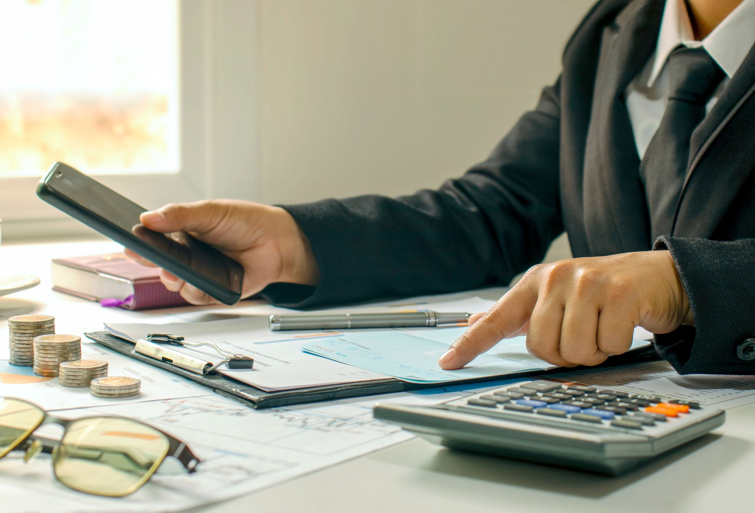 Person in a suit using a smartphone and pointing at financial documents on a desk with coins, a calculator, and a pen, illustrating bookkeeping and financial management services for businesses.